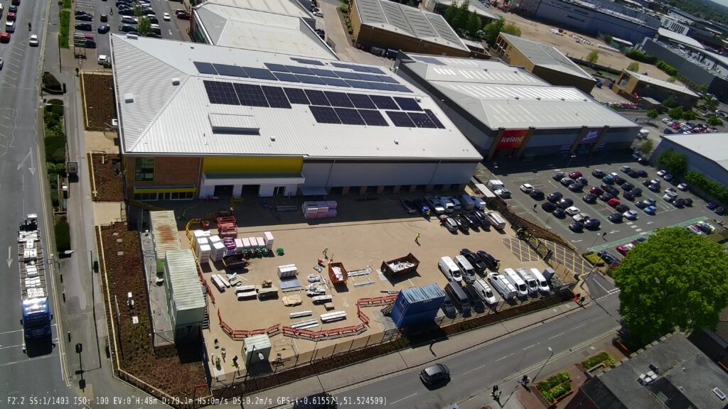 Aerial view of a Big Yellow Self Storage facility. Large building with bright yellow and blue sections, white roofs covered in solar panels. Parking area in front with a few vehicles. Surrounded by roads and green spaces. Shows modern commercial architecture with sustainable energy features.