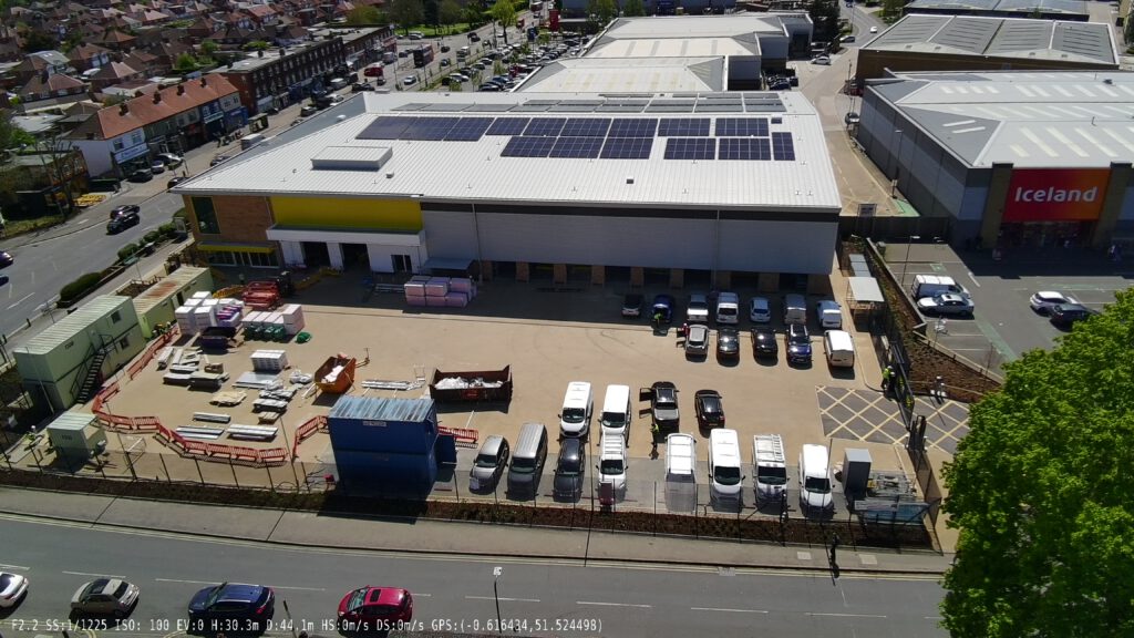 single Photo Aerial view of a Big Yellow Self Storage facility. Large building with bright yellow and blue sections, white roofs covered in solar panels. Parking area in front with a few vehicles. Surrounded by roads and green spaces. Shows modern commercial architecture with sustainable energy features.